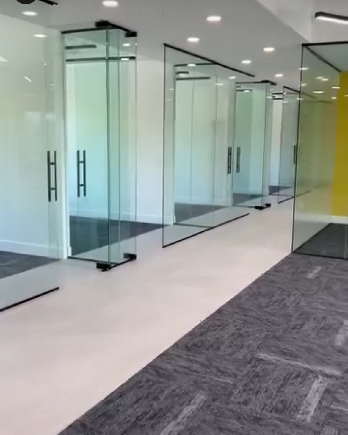 A light central floor path is bordered by dark flooring through a glass-partitioned commercial interior.