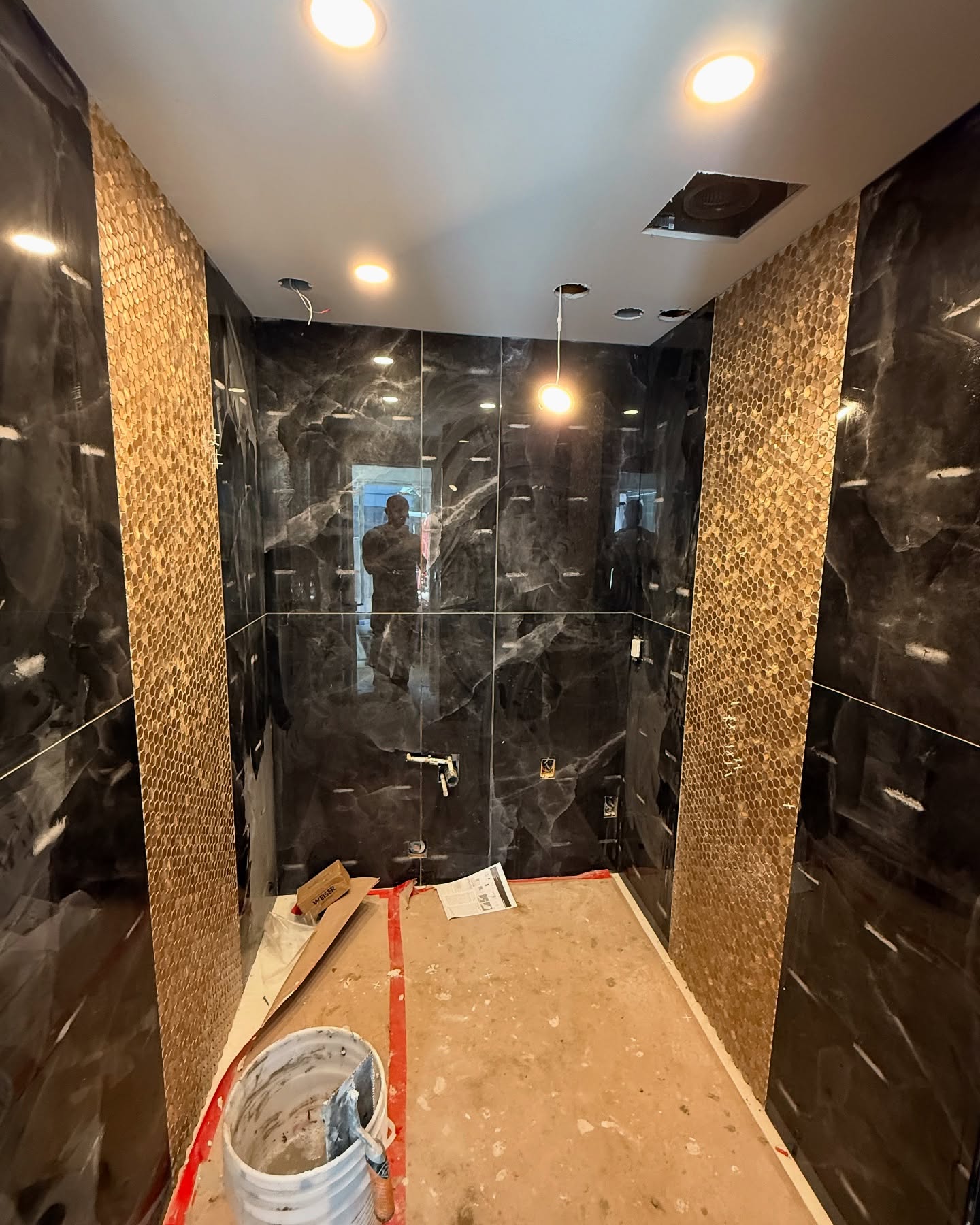 Glossy dark marble-look tile meets vertical bands of gold-toned hexagonal mosaic. Shown during installation.