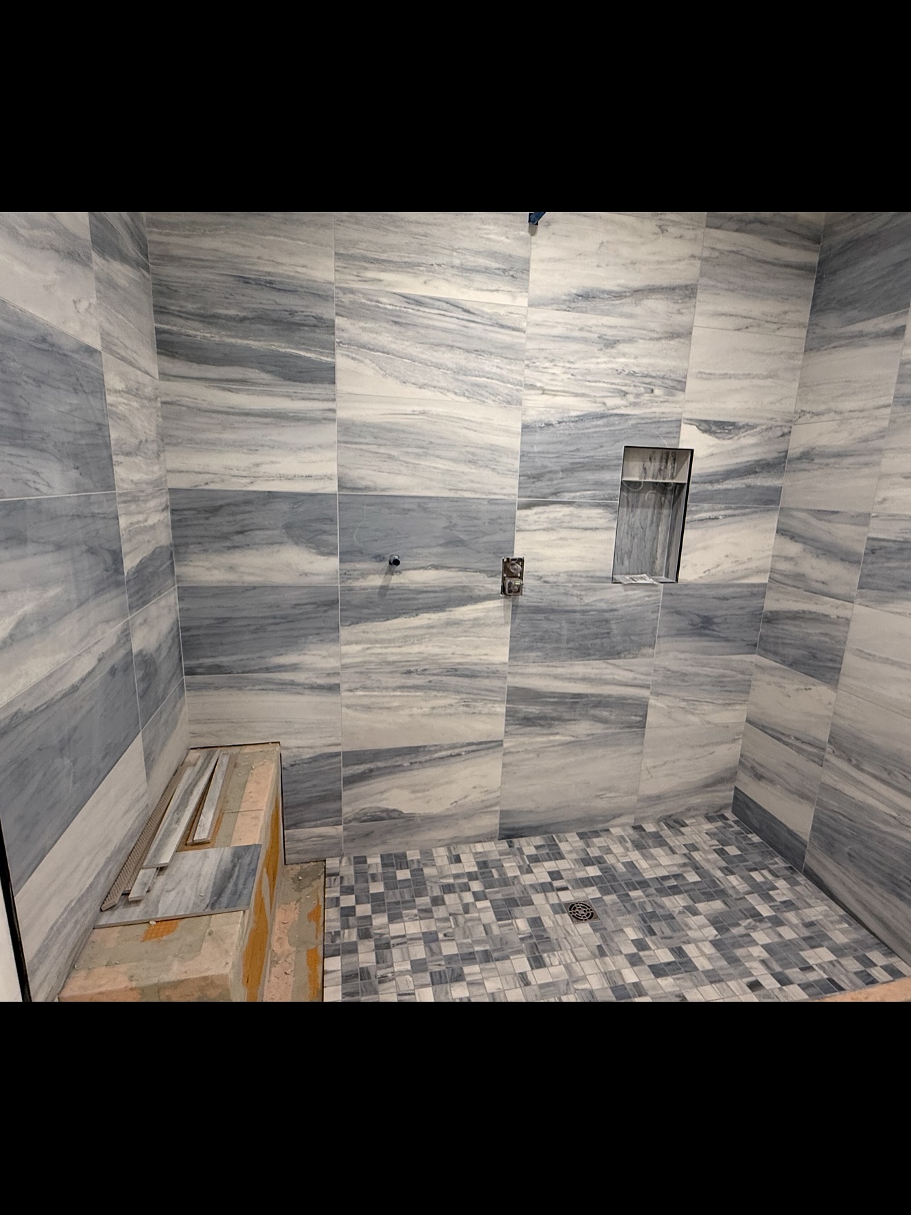 Striated rectangular wall tiles surround a recessed niche above a small square mosaic shower floor. Shown in progress.