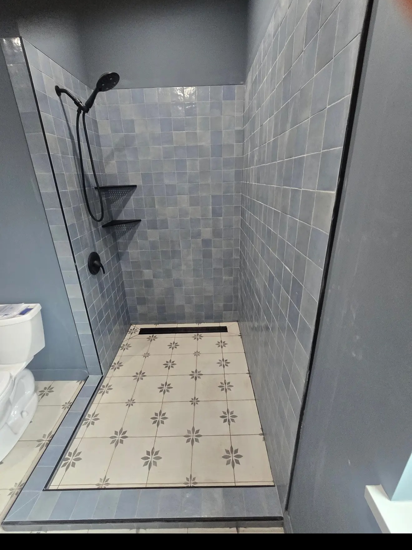 Blue-grey glazed shower walls meet a cream patterned floor, with dark edging and a linear drain.
