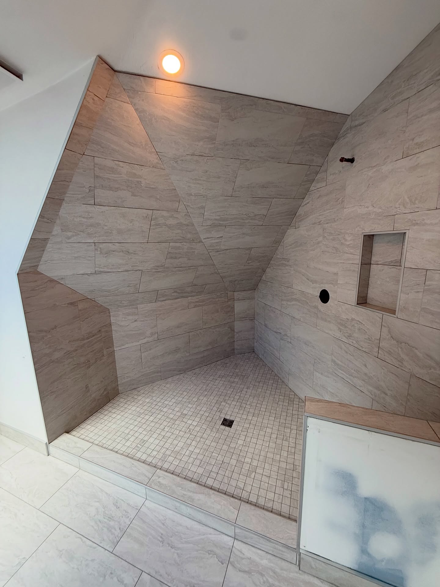 Stone-look tile follows the shower walls and sloped ceiling, with a recessed niche and small-format mosaic base. Shown before final fixtures.