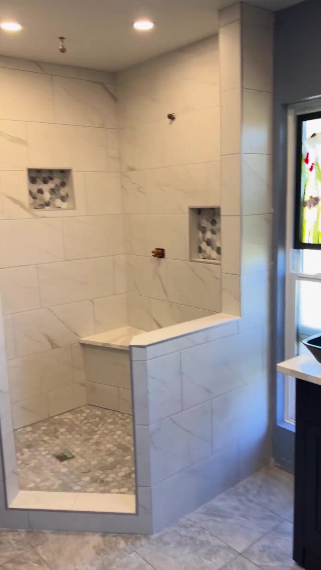 Pale marble-look wall tiles frame mosaic-backed niches and a tiled shower bench above a hexagonal floor. Shown before final fixtures.