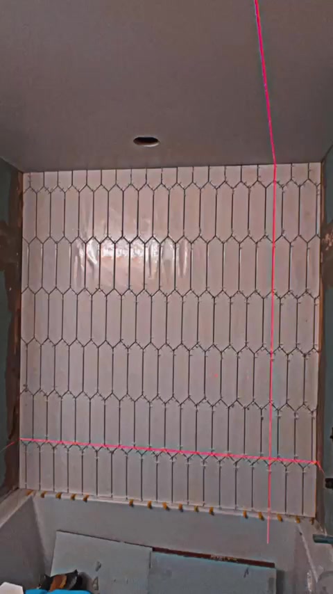 Elongated hexagonal wall tiles are aligned with a visible laser line during installation above the tub.