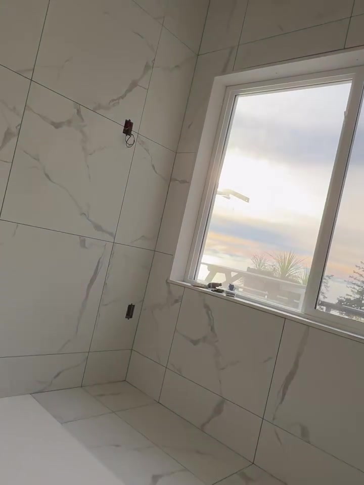 Large pale tiles with grey veining wrap a shower corner and the window reveal. Shown before final fittings.