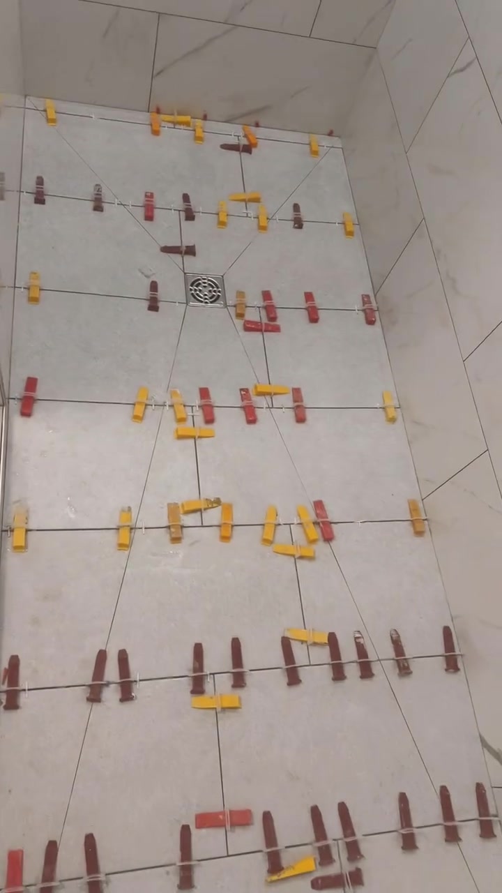 Light rectangular floor tiles are fitted around a square drain, with levelling clips still in place.